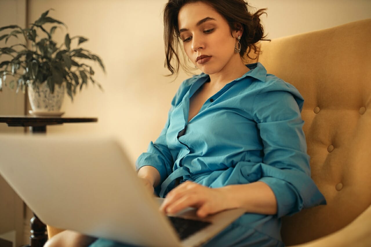 successful-concentrated-young-businesswoman-in-blue-dress-sitting-on-couch-with-portable-computer-on-her-lap